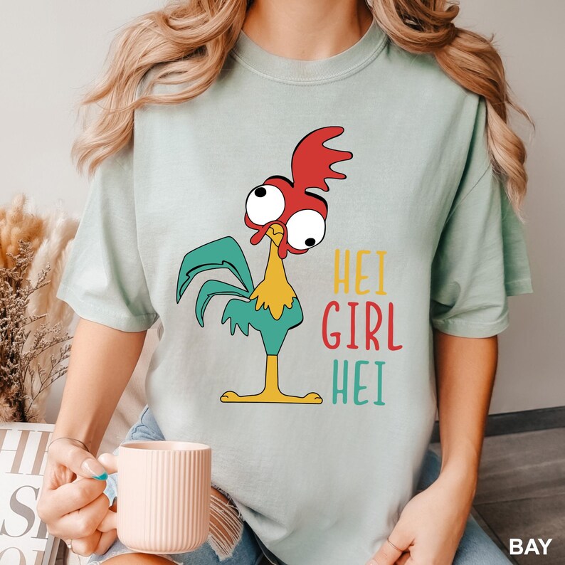 May include: Light green t-shirt featuring a cartoon rooster with red comb, teal wings, yellow chest, and large eyes. The text "HEI GIRL HEI" is printed in yellow, red, and teal. The shirt is a relaxed fit.