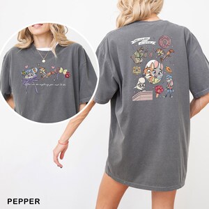 May include: Pepper-coloured t-shirt with a colourful cartoon design. The front of the shirt has a design with the text "You can be anything you wish to be." The back of the shirt has a cartoon design with the text "The Big Donut."
