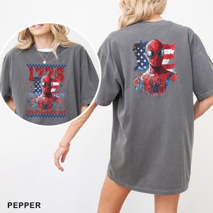 May include: A charcoal gray short-sleeve t-shirt featuring a graphic of Spider-Man against an American flag background. The front design includes the text 1776 UNITED STATES INDEPENDENCE DAY, while the back displays a larger Spider-Man graphic.