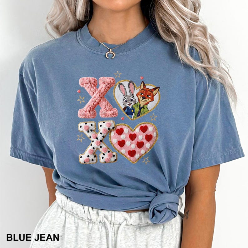 May include: A blue jean colored t-shirt featuring a graphic print of the characters Judy Hopps and Nick Wilde inside a heart, alongside a pink textured X, a white and black polka dot X, and a pink heart with red hearts, accented by small stars.