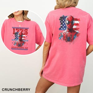 May include: A pink t-shirt featuring a graphic of Spider-Man in front of an American flag. The back displays a large Spider-Man illustration. The front shows the text 1776 UNITED STATES INDEPENDENCE DAY with a checkered border and Spider-Man graphic.