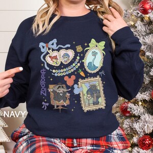 以下が含まれることがあります： ネイビーブルーのスウェットシャツ。ズートピアのキャラクターの額入りイラストが特徴です。デザインには、ニック・ワイルド、ジュディ・ホップス、その他のキャラクターが含まれています。「Zootopia」の文字がスウェットシャツの左側に縦にプリントされています。