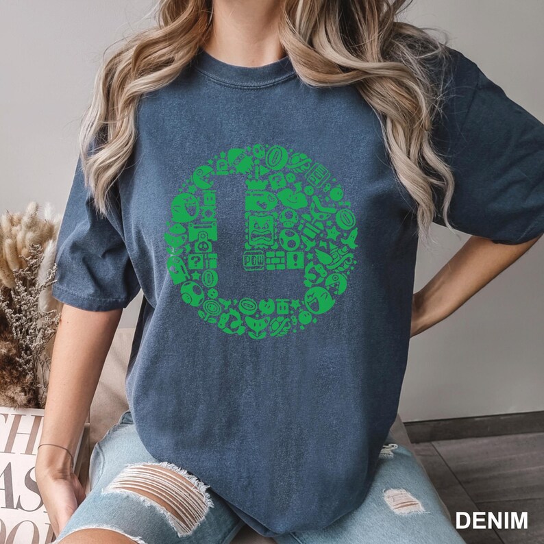 May include: Denim blue t-shirt featuring a green graphic of the letter "L" filled with various video game icons. The shirt has short sleeves and a relaxed fit. The word "DENIM" is printed in white at the bottom right corner.