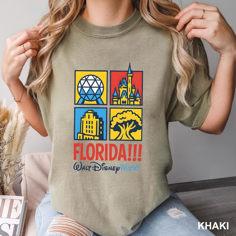 Może przedstawiać: Khaki koszulka z czterema kolorowymi kwadratami z ikonami Disney World. Słowo "FLORIDA!!!" jest wydrukowane na czerwono, a pod nim "Walt Disney World" na niebiesko. Koszulka jest wykonana z miękkiego materiału.