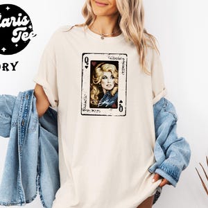 May include: An ivory t-shirt featuring a graphic print of a playing card with a portrait of a blonde woman in a denim jacket. The card design includes the letter Q in the corners and a signature at the bottom. The shirt has short sleeves.