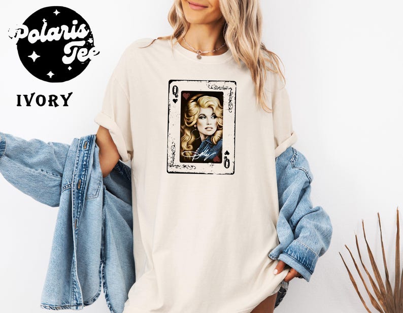 May include: An ivory t-shirt featuring a graphic print of a playing card with a portrait of a blonde woman in a denim jacket. The card design includes the letter Q in the corners and a signature at the bottom. The shirt has short sleeves.