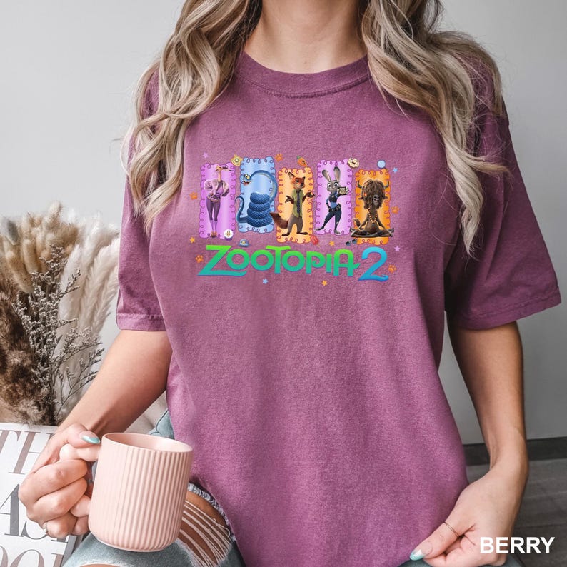 K&ouml;nnte beinhalten: Beerenfarbenes T-Shirt mit dem gr&uuml;nen Text "Zootopia 2" und Illustrationen von Zeichentrickfiguren. Das Shirt hat einen Rundhalsausschnitt und kurze &Auml;rmel. Das Wort "BERRY" ist unten rechts in Wei&szlig; aufgedruckt.