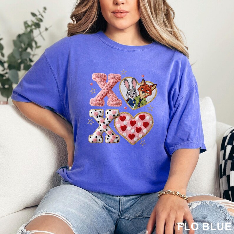 May include: A periwinkle blue short-sleeve t-shirt featuring a graphic print of the letters X and O, a heart containing a rabbit and a fox, and a heart filled with small red hearts. The design includes gold outlines and small star accents.