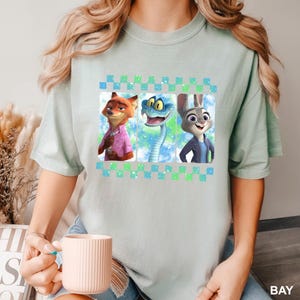 May include: A light green t-shirt featuring a graphic of three cartoon characters: a fox, a snake, and a rabbit. The characters are set against a colorful background within a checkered border. The shirt is a light green color.