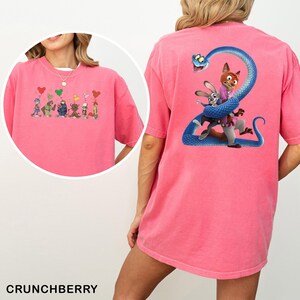 Op de afbeelding: Een roze t-shirt met cartoonontwerpen op de voor- en achterkant. Het ontwerp aan de voorkant toont cartoonfiguren met hartvormige ballonnen. Het ontwerp aan de achterkant toont een blauwe slang die zich om cartoonfiguren wikkelt. Het woord "CRUNCHBERRY" staat onderaan gedrukt.