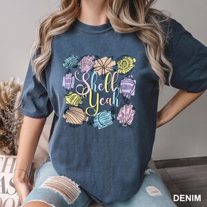 May include: A denim blue t-shirt with a colorful seashell design and the words "Shell Yeah" in a script font. The seashells are in various colors, including yellow, pink, blue, and green. The shirt is a casual style.