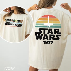 May include: Ivory t-shirt with "DISNEY CREW" text and cartoon characters on the front. The back has a retro sunset design with an X-wing fighter and "STAR WARS 1977". The shirt is a cream colour.