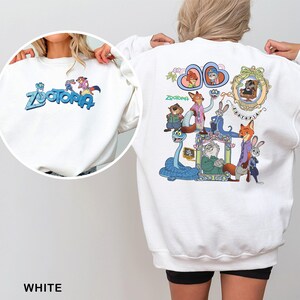 Puede incluir: Sudadera blanca con la palabra "Zootopia" y personajes de dibujos animados en la parte delantera y trasera. El dise&ntilde;o trasero incluye varios personajes de animales y el logotipo de Zootopia. La sudadera es de cuello redondo.