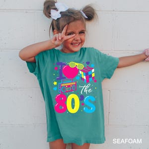 May include: A seafoam green t-shirt featuring a colorful graphic print that reads I Love the 80s. The design includes a bright pink heart, yellow sunglasses, a multi-colored cube puzzle, a boombox, and various small stars and hearts in vibrant colors.