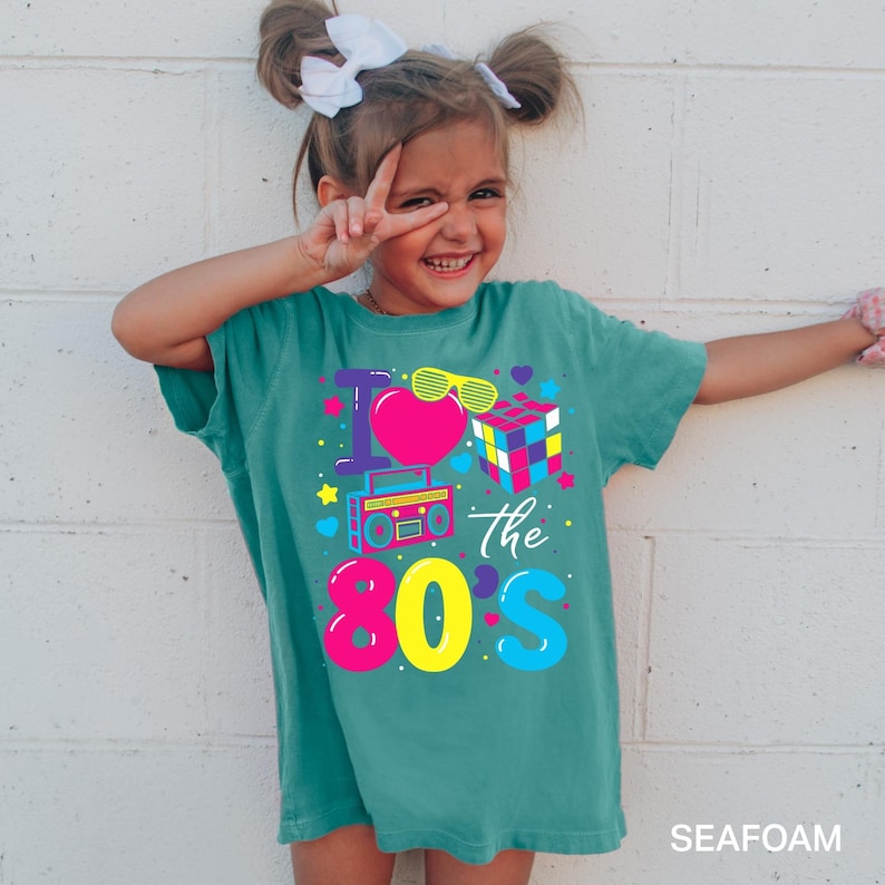 May include: A seafoam green t-shirt featuring a colorful graphic print that reads I Love the 80s. The design includes a bright pink heart, yellow sunglasses, a multi-colored cube puzzle, a boombox, and various small stars and hearts in vibrant colors.