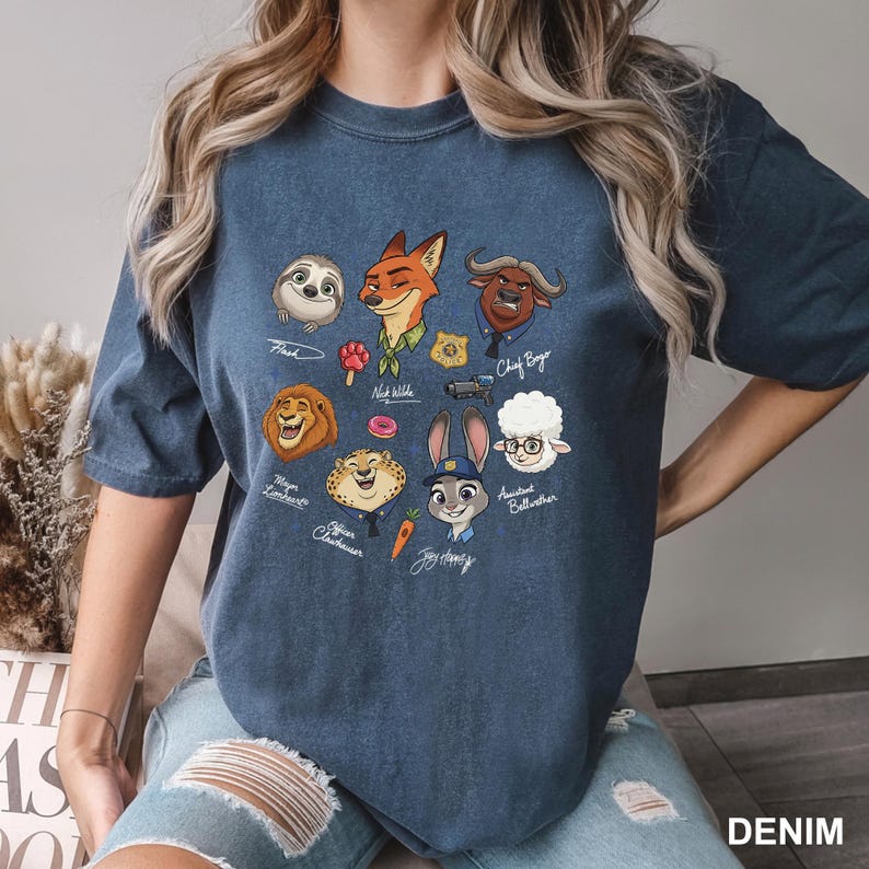 K&ouml;nnte beinhalten: Denimblaues T-Shirt mit Cartoon-Charakterportr&auml;ts aus Zootopia. Das Shirt zeigt Flash, Nick Wilde, Chief Bogo, Mayor Lionheart, Officer Clawhauser, Judy Hopps und andere. Das Wort "DENIM" ist unten rechts aufgedruckt.