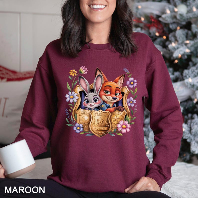 May include: Maroon sweatshirt featuring a cartoon image of Judy Hopps and Nick Wilde from Zootopia, framed by a gold police badge and floral accents. The word "MAROON" is printed at the bottom.