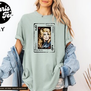 May include: A sage green short-sleeve t-shirt featuring a graphic print of a playing card with the Queen of Hearts. The card displays a portrait of a blonde woman with a signature below, framed by a black border with ornate corner details.