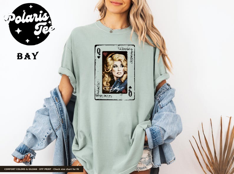 May include: A sage green short-sleeve t-shirt featuring a graphic print of a playing card with the Queen of Hearts. The card displays a portrait of a blonde woman with a signature below, framed by a black border with ornate corner details.