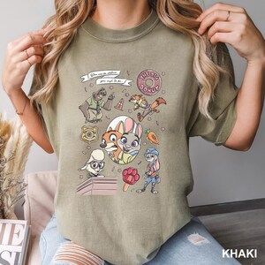 Op de afbeelding: Khaki T-shirt met een kleurrijke afbeelding van Zootopia-personages, waaronder Judy Hopps en Nick Wilde. Het ontwerp bevat de tekst "You can be whatever you wish to be" en "The Big Donut".