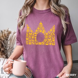 May include: Berry-colored t-shirt featuring a yellow crown design filled with video game icons. The shirt has short sleeves and a relaxed fit. The word "BERRY" is printed in white at the bottom right corner of the image.