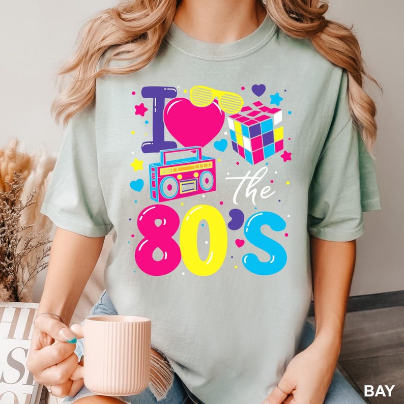 K&ouml;nnte beinhalten: Hellgr&uuml;nes T-Shirt mit einem farbenfrohen Aufdruck "I love the 80's". Das Design umfasst ein rosa Herz, einen Rubik's Cube, einen Ghettoblaster und andere Elemente im 80er-Jahre-Stil.