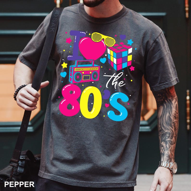 May include: A charcoal gray short-sleeve t-shirt featuring a vibrant graphic print that reads I Love the 80s. The design includes a bright pink heart, yellow shutter shades, a colorful puzzle cube, a blue boombox, and various small stars and hearts.