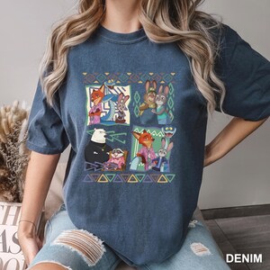 K&ouml;nnte beinhalten: Denimblaues T-Shirt mit einem farbenfrohen Grafikdesign von Charakteren aus einem beliebten Zeichentrickfilm. Das Design zeigt Bilder eines Fuchses, eines Kaninchens und anderer Tierfiguren in verschiedenen Posen. Das Wort "DENIM" ist unten rechts in Wei&szlig; gedruckt.