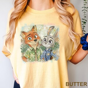 May include: Yellow t-shirt featuring a distressed graphic of Nick Wilde and Judy Hopps from Zootopia. Nick is wearing a green shirt and tie, while Judy is in a police uniform. The word "BUTTER" is printed at the bottom of the shirt.
