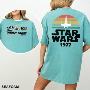 May include: Seafoam green t-shirt with a Disney Crew graphic on the front and a Star Wars design on the back. The Star Wars graphic includes a white X-wing fighter, a sunset, and the text "STAR WARS 1977".
