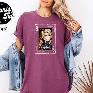 May include: A berry-colored short-sleeve t-shirt featuring a graphic print of a playing card with the letter Q and a heart symbol. The card displays a portrait of a blonde woman with the signature Dolly written in white script across the bottom.
