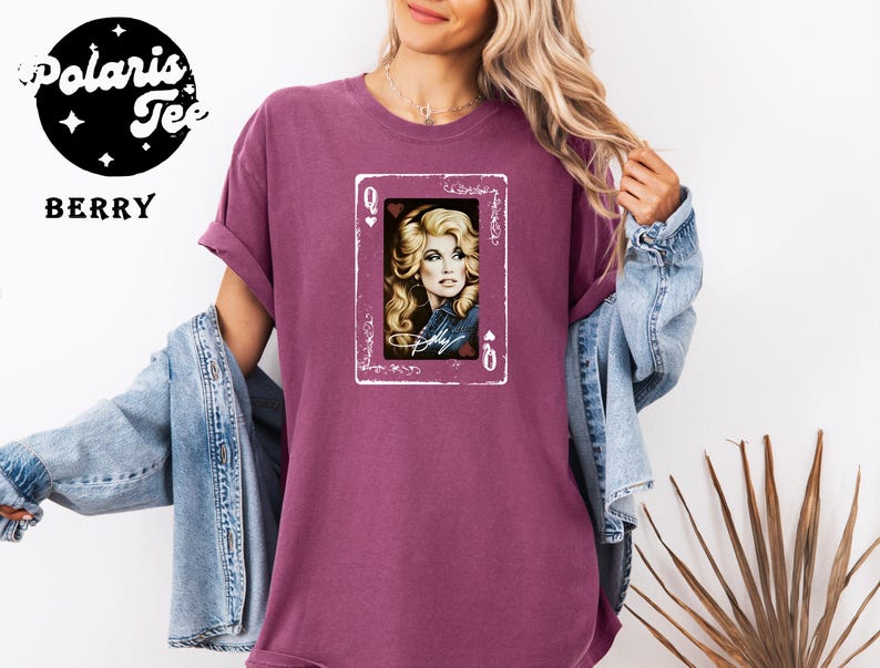 May include: A berry-colored short-sleeve t-shirt featuring a graphic print of a playing card with the letter Q and a heart symbol. The card displays a portrait of a blonde woman with the signature Dolly written in white script across the bottom.