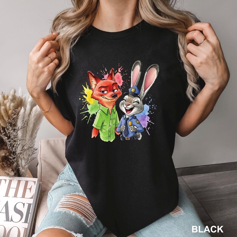 May include: Black t-shirt featuring a colorful graphic of Nick Wilde and Judy Hopps from Zootopia. Nick is a red fox wearing a green shirt, and Judy is a gray rabbit in a police uniform. The word "BLACK" is printed at the bottom.