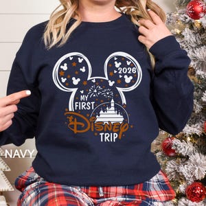 May include: Navy blue sweatshirt featuring a Mickey Mouse head design with the text "My First Disney Trip" and the year "2026". The design includes stars, fireworks, and a castle. The word "NAVY" is printed on the bottom left.