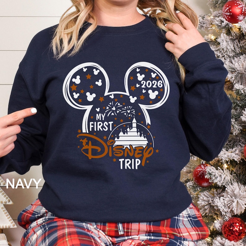 May include: Navy blue sweatshirt featuring a Mickey Mouse head design with the text "My First Disney Trip" and the year "2026". The design includes stars, fireworks, and a castle. The word "NAVY" is printed on the bottom left.