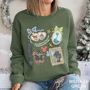 以下が含まれることがあります： Zootopiaのキャラクターが額縁とハート型のロケットに入った、ミリタリーグリーンのスウェットシャツ。デザインにはニック・ワイルド、ジュディ・ホップス、その他のキャラクターが含まれています。「Zootopia」の文字が紫で縦書きされています。