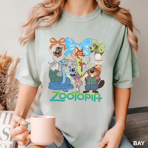 K&ouml;nnte beinhalten: Hellgr&uuml;nes T-Shirt mit einem farbenfrohen Grafikdesign von Charakteren aus dem Film "Zoomania". Das Design umfasst eine blaue Schleife, einen goldenen Rahmen und das Wort "Zootopia" in Gr&uuml;n und Blau.
