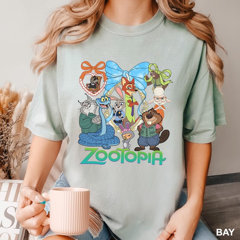 K&ouml;nnte beinhalten: Hellgr&uuml;nes T-Shirt mit einem farbenfrohen Grafikdesign von Charakteren aus dem Film "Zoomania". Das Design umfasst eine blaue Schleife, einen goldenen Rahmen und das Wort "Zootopia" in Gr&uuml;n und Blau.