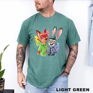 May include: Light green t-shirt featuring a colorful graphic of a fox and a rabbit, both smiling. The fox is wearing a green shirt, and the rabbit is in a police uniform. The words "LIGHT GREEN" are at the bottom of the shirt.