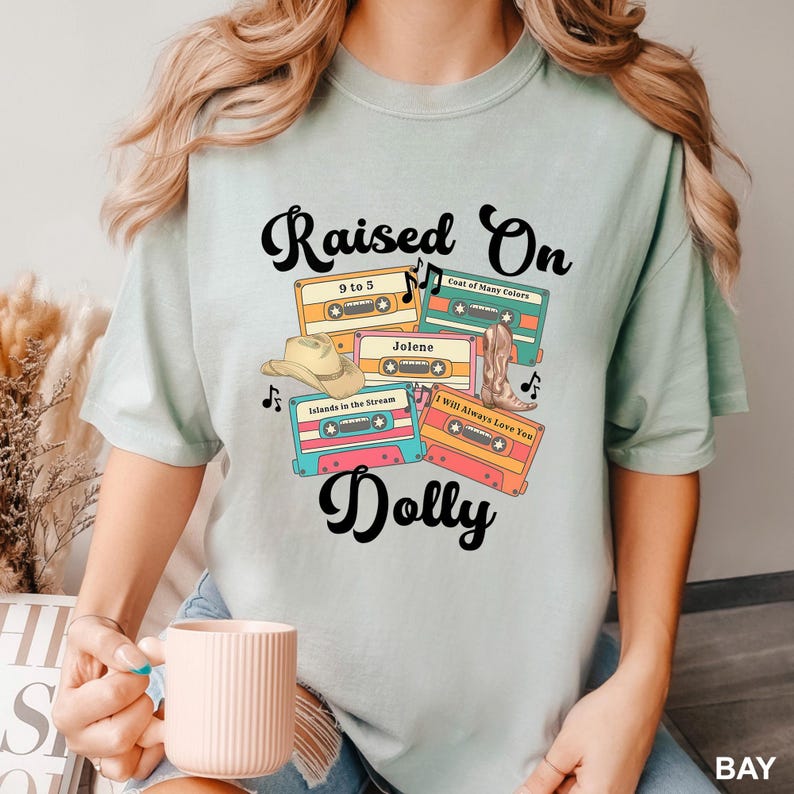 May include: A light green t-shirt featuring a graphic print of four colorful cassette tapes labeled 9 to 5, Coat of Many Colors, Jolene, Islands in the Stream, and I Will Always Love You, with a cowboy hat, a boot, and the text Raised On Dolly.