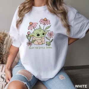 Puede incluir: Camiseta blanca con un personaje de dibujos animados, flores rosas y una peque&ntilde;a rana verde. El texto "Enjoy the Little Things" est&aacute; impreso debajo. La camiseta es de estilo casual.
