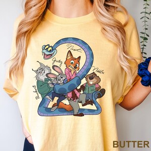 Peut inclure: T-shirt jaune beurre avec un dessin anim&eacute; de personnages de Zootopie. Le design comprend Nick Wilde, Judy Hopps et d'autres personnages, avec le mot "Butter" imprim&eacute; en bas.