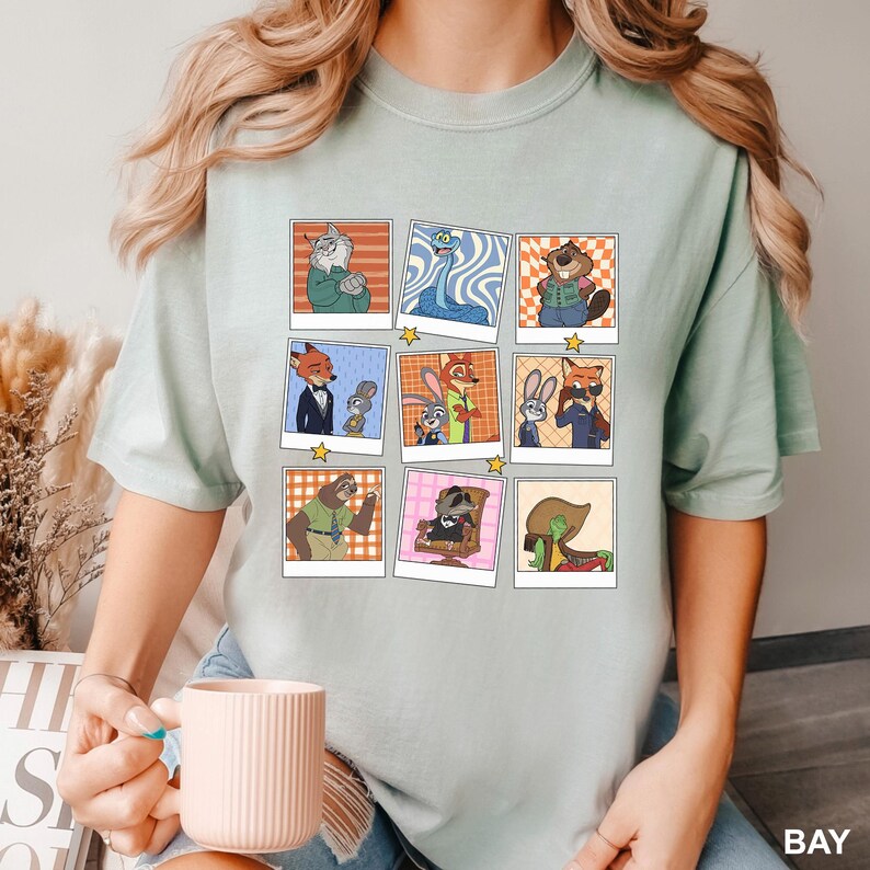 May include: Light green t-shirt featuring a grid of cartoon character portraits in polaroid-style frames. The characters include a fox in a suit, a sloth, and a raccoon. The word "BAY" is printed on the bottom right.