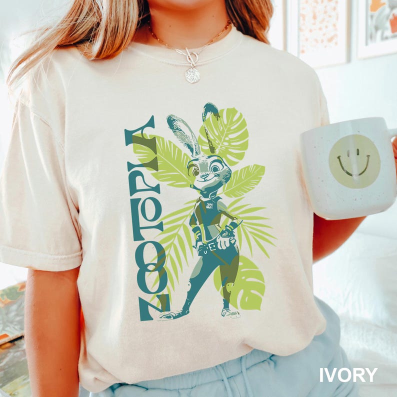 May include: Ivory-colored t-shirt featuring a Zootopia graphic. The design includes the word "Zootopia" in teal, a cartoon rabbit character, and green tropical leaves. A white mug with a smiley face is also visible.