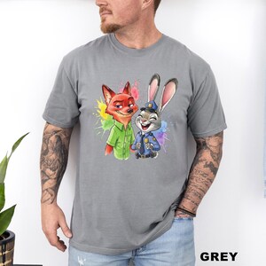 May include: Grey t-shirt featuring a colorful graphic of Nick Wilde the fox and Judy Hopps the rabbit from Zootopia. Nick is wearing a green shirt, and Judy is in a police uniform. The word "GREY" is printed in the bottom right corner.