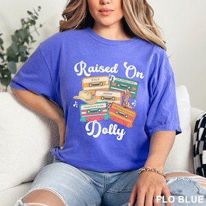 May include: A bright blue short-sleeve t-shirt featuring the white text "Raised On Dolly" and a graphic of four colorful cassette tapes labeled "9 to 5," "Coat of Many Colors," "Jolene," "Islands in the Stream," and "I Will Always Love You" with a cowboy hat and boot.