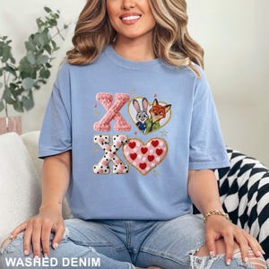 May include: A light blue t-shirt featuring a graphic print of the letters X and O in a textured pink knit pattern, alongside a heart containing cartoon characters Judy Hopps and Nick Wilde, and a second heart filled with small red hearts.
