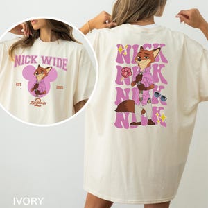 May include: Ivory t-shirt with a cartoon fox character from Zootopia. The front has "NICK WIDE" text and a character graphic. The back displays the character with "NICK" repeated vertically, along with sunglasses and a popsicle.