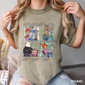 K&ouml;nnte beinhalten: Khakifarbenes T-Shirt mit einem farbenfrohen Grafikmuster von animierten Tierfiguren in einem quadratischen Muster. Das Design umfasst einen Fuchs, ein Kaninchen, einen B&auml;ren und andere Charaktere. Das Wort "KHAKI" ist unten rechts aufgedruckt.