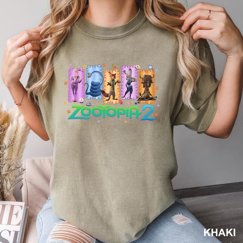 K&ouml;nnte beinhalten: Khaki-farbenes T-Shirt mit dem Text "Zootopia 2" in Gr&uuml;n und einer bunten Grafik von Charakteren aus dem Animationsfilm. Die Grafik enth&auml;lt Bilder eines Geparden, einer Schlange, eines Fuchses, eines Kaninchens und eines Yaks.